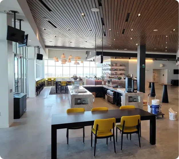 Modern cafeteria with a sleek design featuring a long black table with yellow chairs, a bar area, TV screens, and large windows letting in natural light. Expertly designed by commercial electrical contractors to ensure an efficient and vibrant atmosphere.