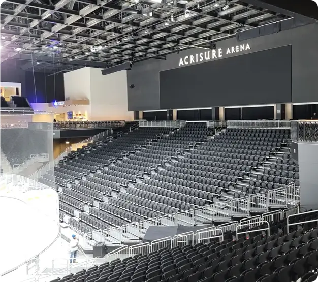 Empty, well-lit, indoor arena seating area with black chairs and a large blank screen displaying the text "Acrisure Arena" above. A worker from the commercial electrical contractors is visible on the left side near the ice rink.