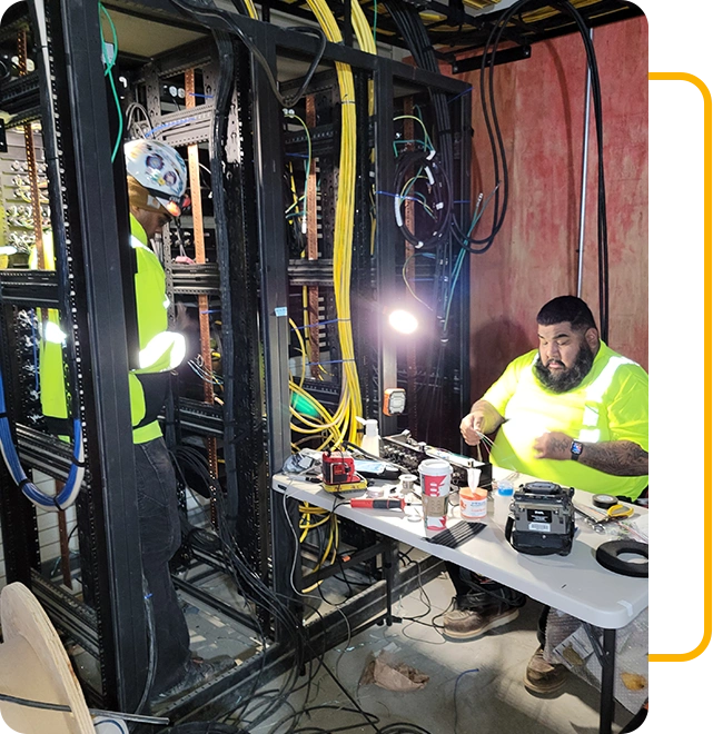 Two workers in yellow shirts handle networking equipment in a server room, one standing working on cables while the other sits at a table with tools and a light, showcasing professional electrical services at its best.